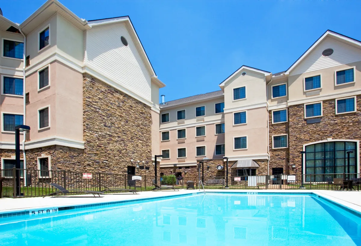 A vibrant outdoor pool is surrounded by a stone facade and the building's attractive architecture. Guests can be seen swimming, sunbathing on loungers, and relaxing by the poolside, all under the open sky. The patio area nearby hints at gatherings and social moments, while the scene captures a refreshing, lively mood ideal for warm weather leisure.