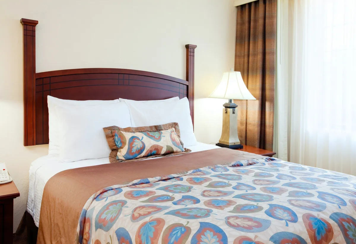 A bright, comfortable bedroom featuring a king-sized bed adorned with a decorative pillow and warm quilt. Natural light filters through the curtains, and a bedside lamp provides additional illumination, creating an inviting environment for relaxation and restful sleep.