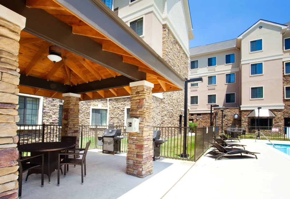 A welcoming exterior view featuring a covered patio with comfortable outdoor seating, a BBQ grill area, and the pool in the background. The scene is set for outdoor barbecuing, casual relaxation, and social gatherings, capturing the communal and inviting nature of the space.