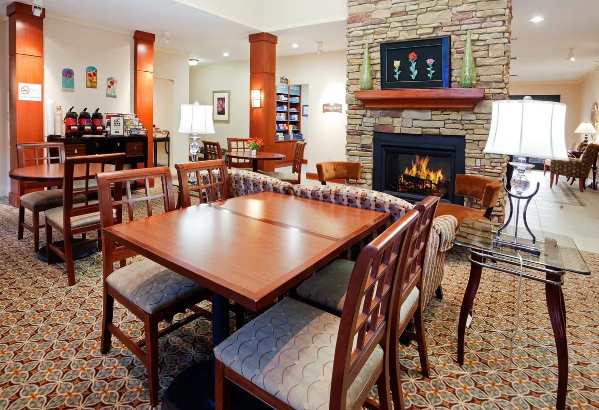 A cozy common area featuring a fireplace, wooden tables, comfortable seating, and artwork on the walls. The space is arranged to invite social gatherings, conversations over coffee, or quiet relaxation by the fire.