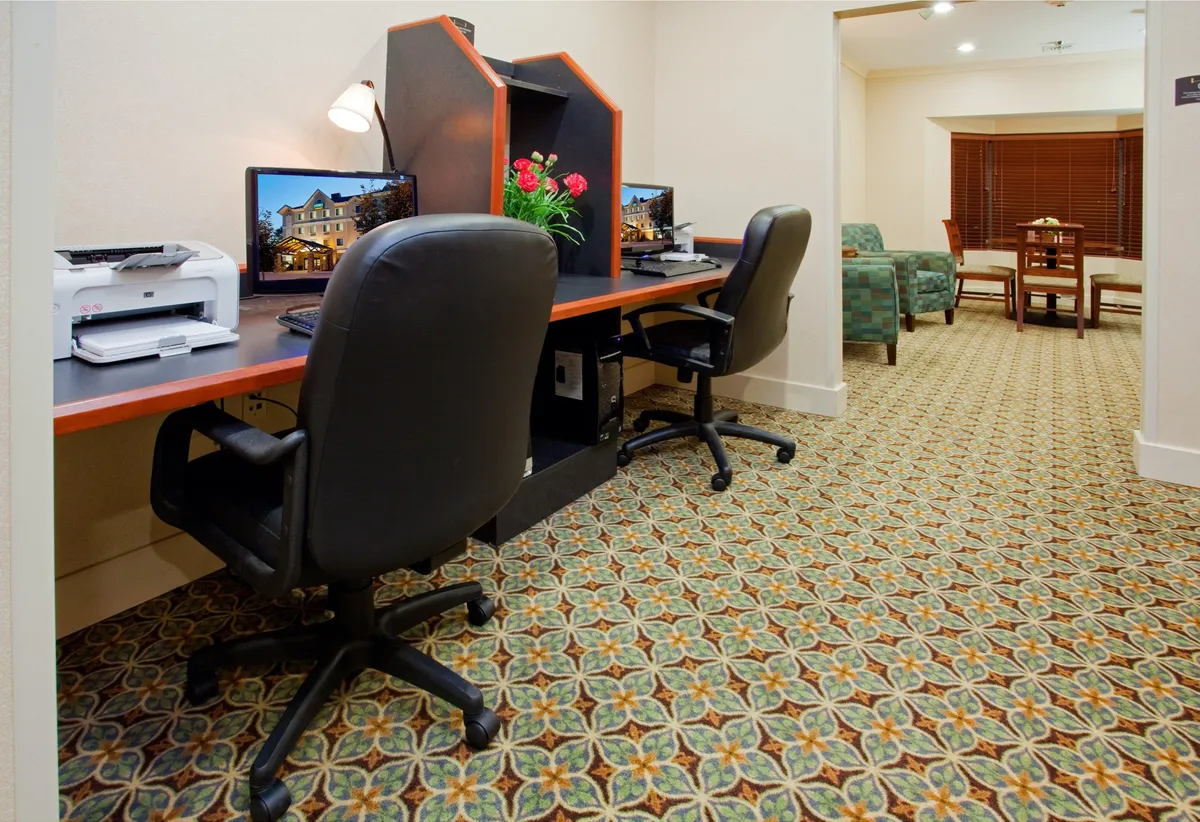 A comfortable business center setup is shown here, with two computer desks, a printer, a green plant, and soft carpeting. The space invites productivity while offering a relaxed atmosphere for work or quick tasks.