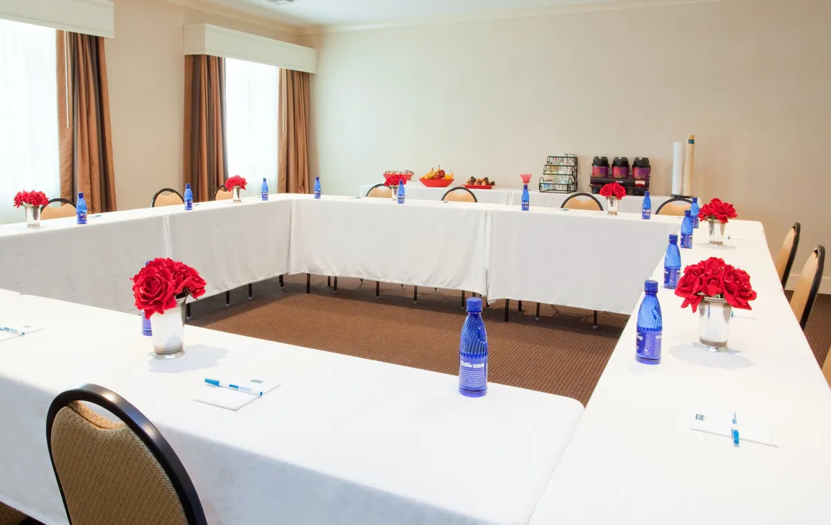 A well-appointed meeting room featuring a long rectangular table, vases of red roses, blue water bottles, and snacks on a side table. The setting is ideal for business meetings, workshops, or collaborative gatherings.